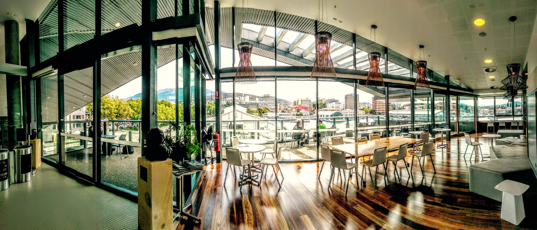 A wide view of a bright room with wooden floors, tables and chairs, and tall windows looking out over Hobart's harbour and a mountain.
