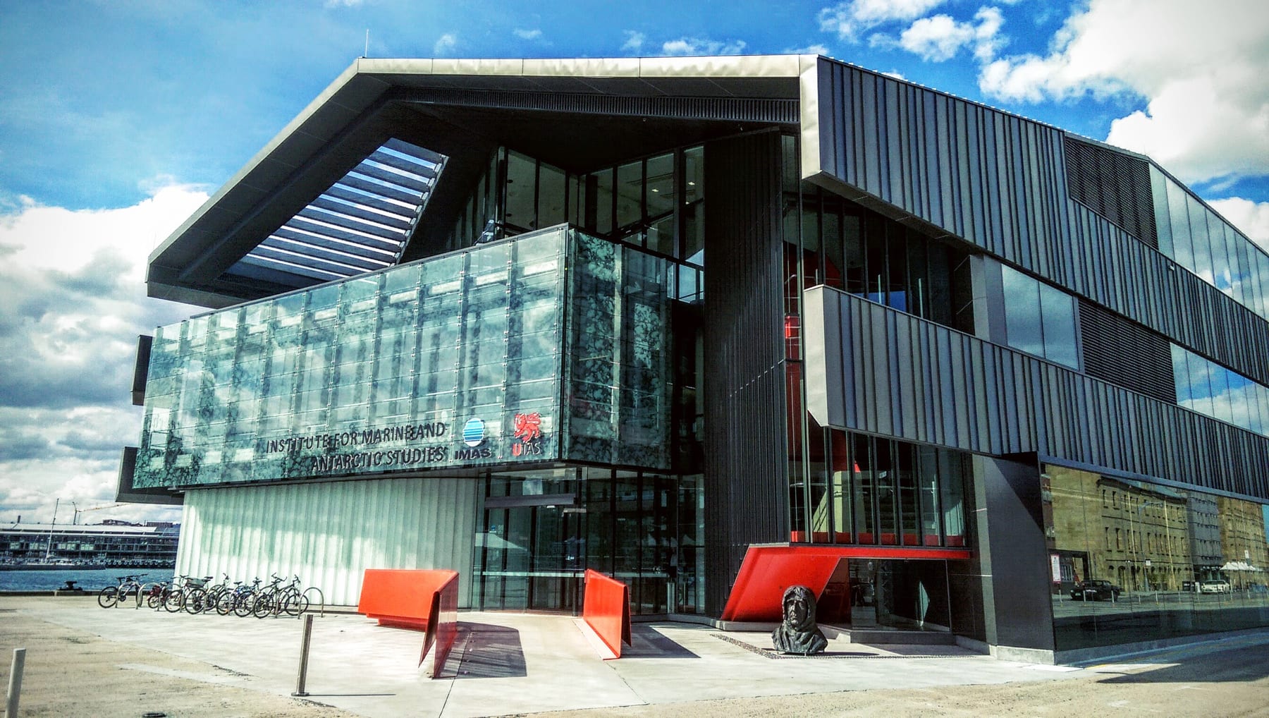 A modern building of glass and dark metal with the sign 'Institute for Marine and Antarctic Studies', red benches and bicycles in front, under a blue sky.