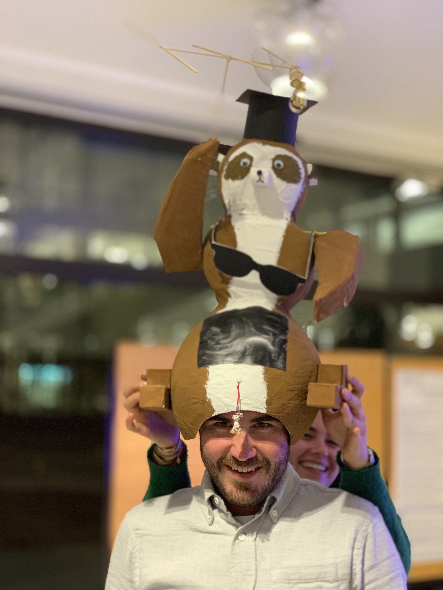 A smiling man wearing a large, home-made paper meerkat hat with a graduation cap on top, held in place by a laughing woman standing behind him.